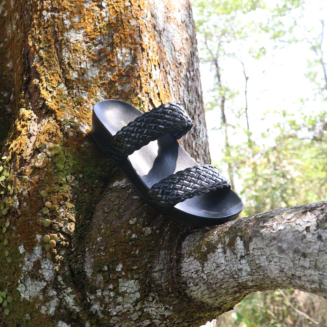 A single black twin-band flatform sandal with a woven texture rests diagonally on the trunk of a large mossy tree in an outdoor nature setting, with sunlight filtering through green foliage in the background.