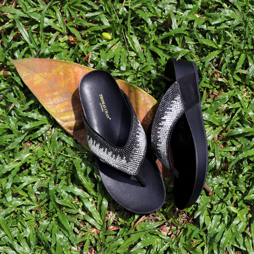 Suede rhinestone flip-flops in black from Thomas Chan's LiteStep Collection placed on fresh green grass with dried leaves, showcasing the shimmering rhinestone straps and lightweight sole.