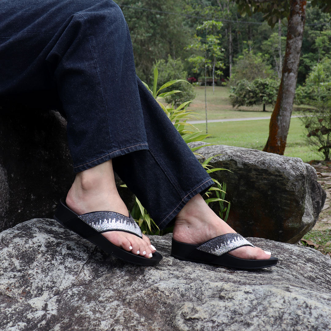 Model seated on a stone outdoors wearing Thomas Chan’s black suede rhinestone flip-flops, highlighting the relaxed style, glittering straps, and comfy flatform sole.
