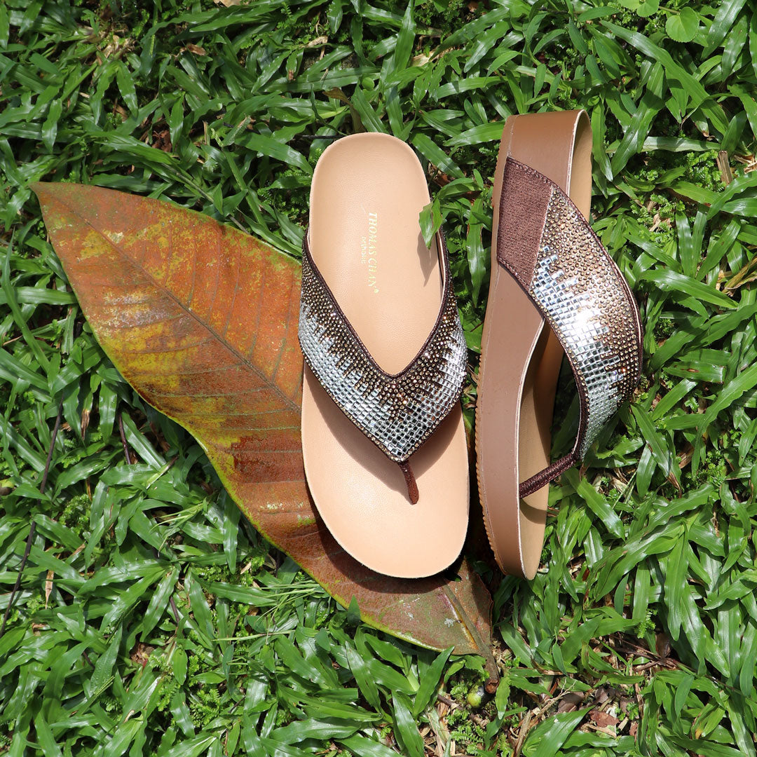 Suede rhinestone flip-flops in bronze from Thomas Chan's LiteStep Collection placed on fresh green grass with dried leaves, showcasing the shimmering rhinestone straps and lightweight sole.