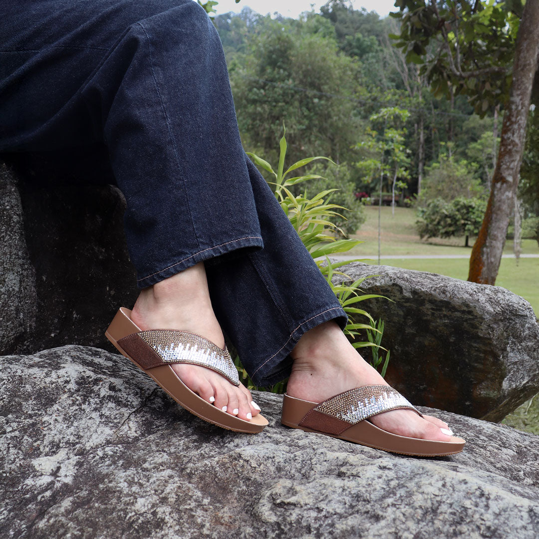 Model seated on a stone outdoors wearing Thomas Chan’s bronze suede rhinestone flip-flops, highlighting the relaxed style, glittering straps, and comfy flatform sole.