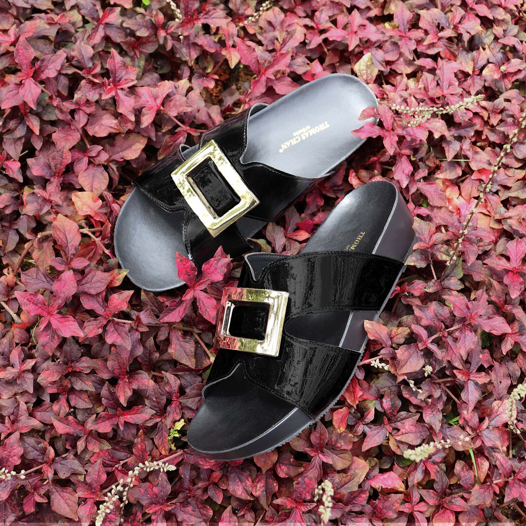 Thomas Chan LiteStep flatform sandals in black patent leather with square gold buckles, laid on vibrant red foliage background.
