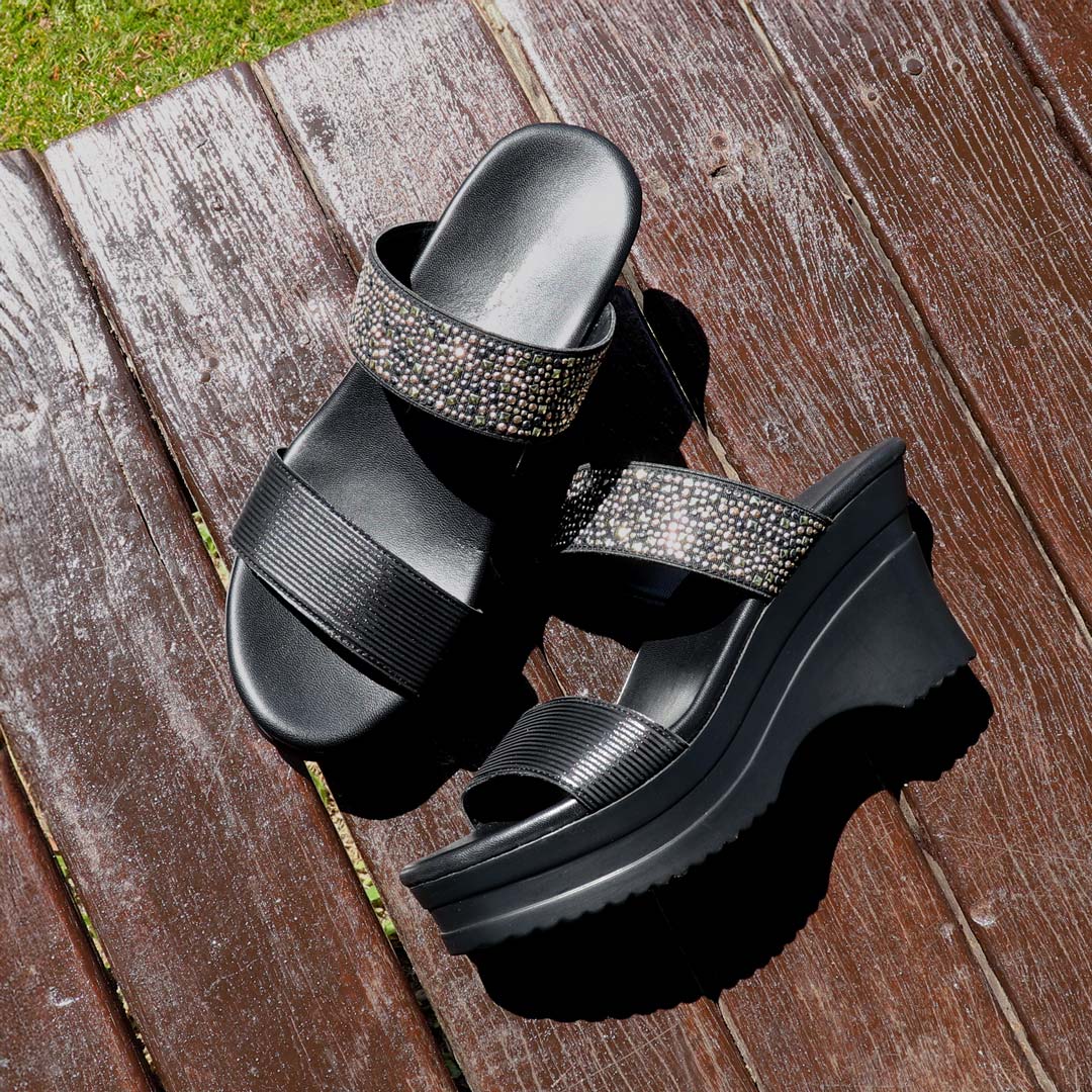 Thomas Chan's Strappy Rhinestone Cushioned Heels in black are elegantly displayed on a rustic wooden bench in an outdoor pavilion setting. The sleek black straps adorned with shimmering rhinestones create a striking contrast against the sunlit wood. Natural sunlight highlights the heels' sophisticated design, while the warm earthy tones of the setting add a touch of rustic charm, emphasizing their elegance and versatility.