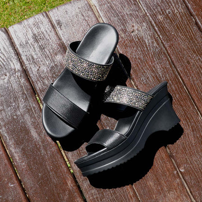 Thomas Chan's Strappy Rhinestone Cushioned Heels in black are elegantly displayed on a rustic wooden bench in an outdoor pavilion setting. The sleek black straps adorned with shimmering rhinestones create a striking contrast against the sunlit wood. Natural sunlight highlights the heels' sophisticated design, while the warm earthy tones of the setting add a touch of rustic charm, emphasizing their elegance and versatility.