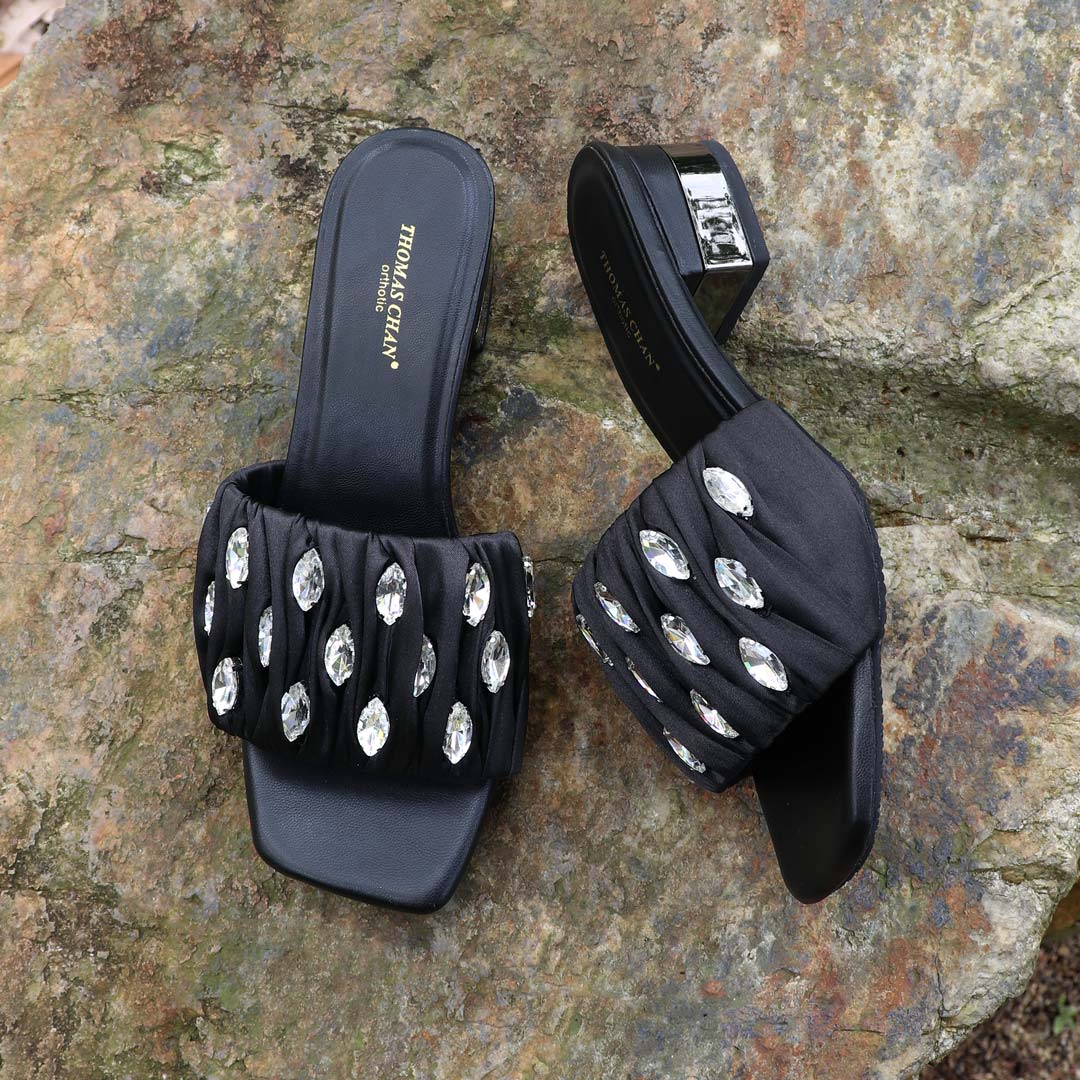 Crystal Lux Satin Evening Heels in black displayed outdoors on a textured stone surface, featuring sparkling crystal embellishments and a glossy block heel — perfect for elegant dinner and evening wear.