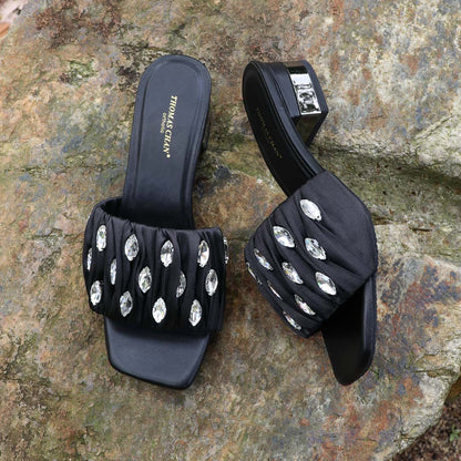 Crystal Lux Satin Evening Heels in black displayed outdoors on a textured stone surface, featuring sparkling crystal embellishments and a glossy block heel — perfect for elegant dinner and evening wear.
