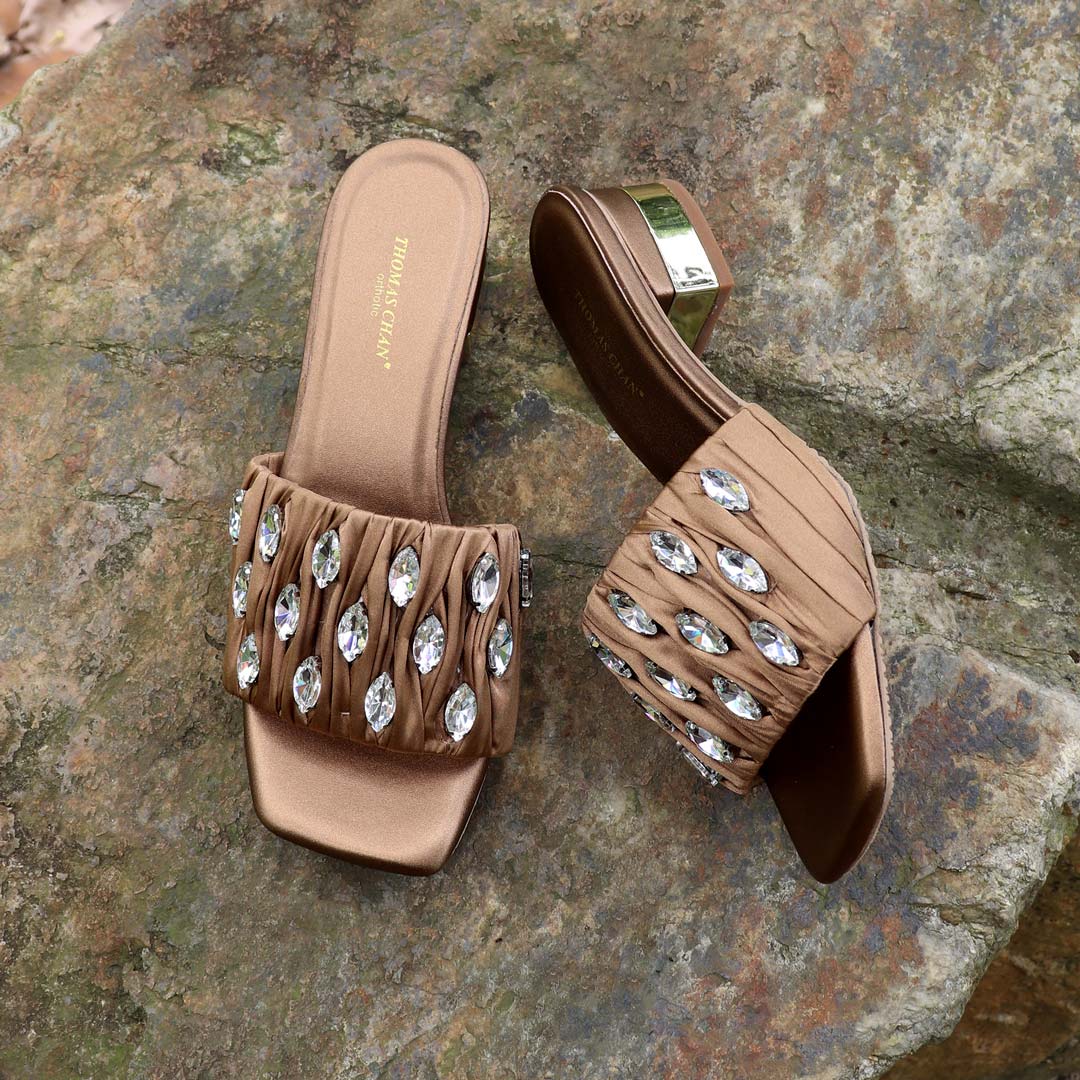 Crystal Lux Satin Evening Heels in bronze displayed outdoors on a textured stone surface, featuring sparkling crystal embellishments and a glossy block heel — perfect for elegant dinner and evening wear.