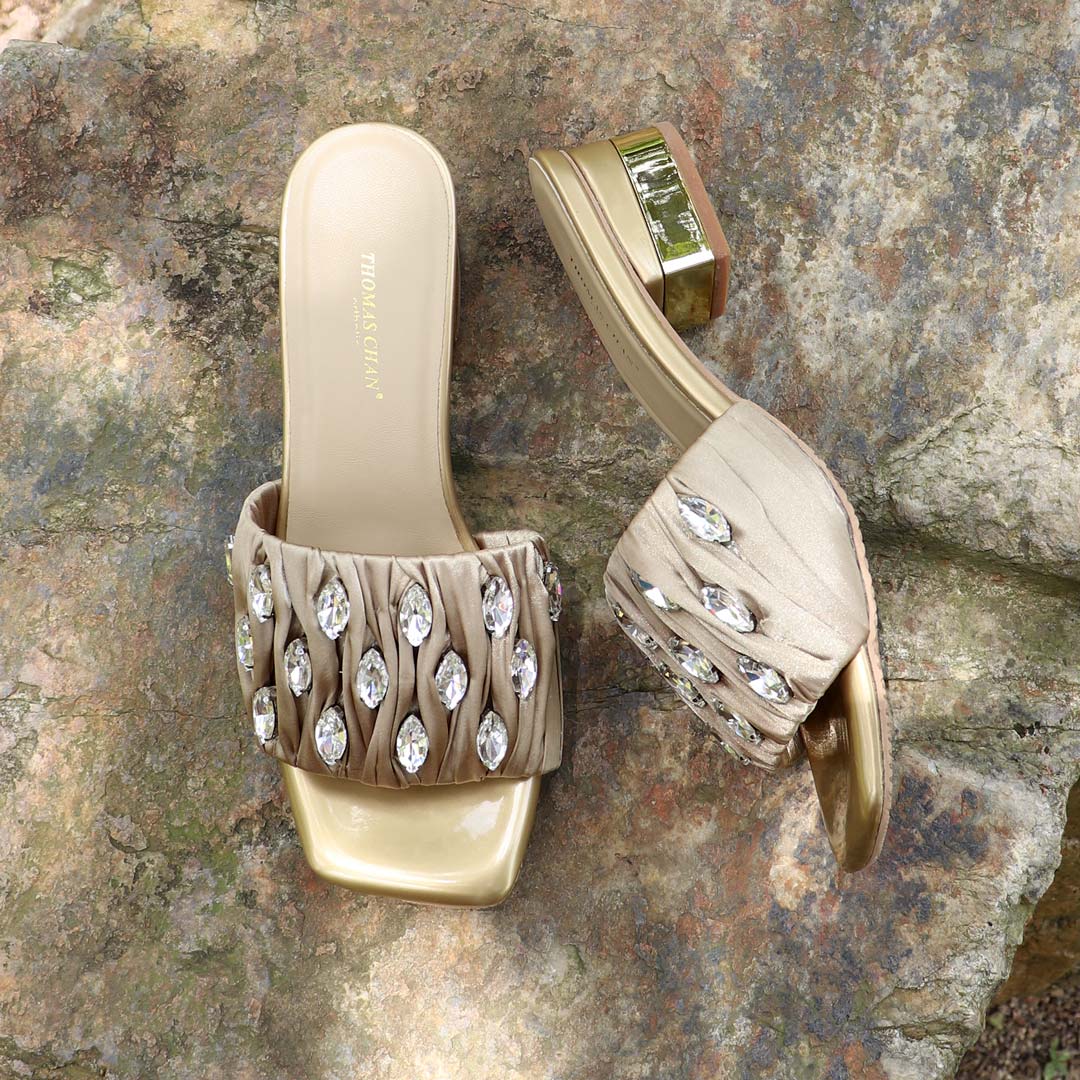 Crystal Lux Satin Evening Heels in gold displayed outdoors on a textured stone surface, featuring sparkling crystal embellishments and a glossy block heel — perfect for elegant dinner and evening wear.