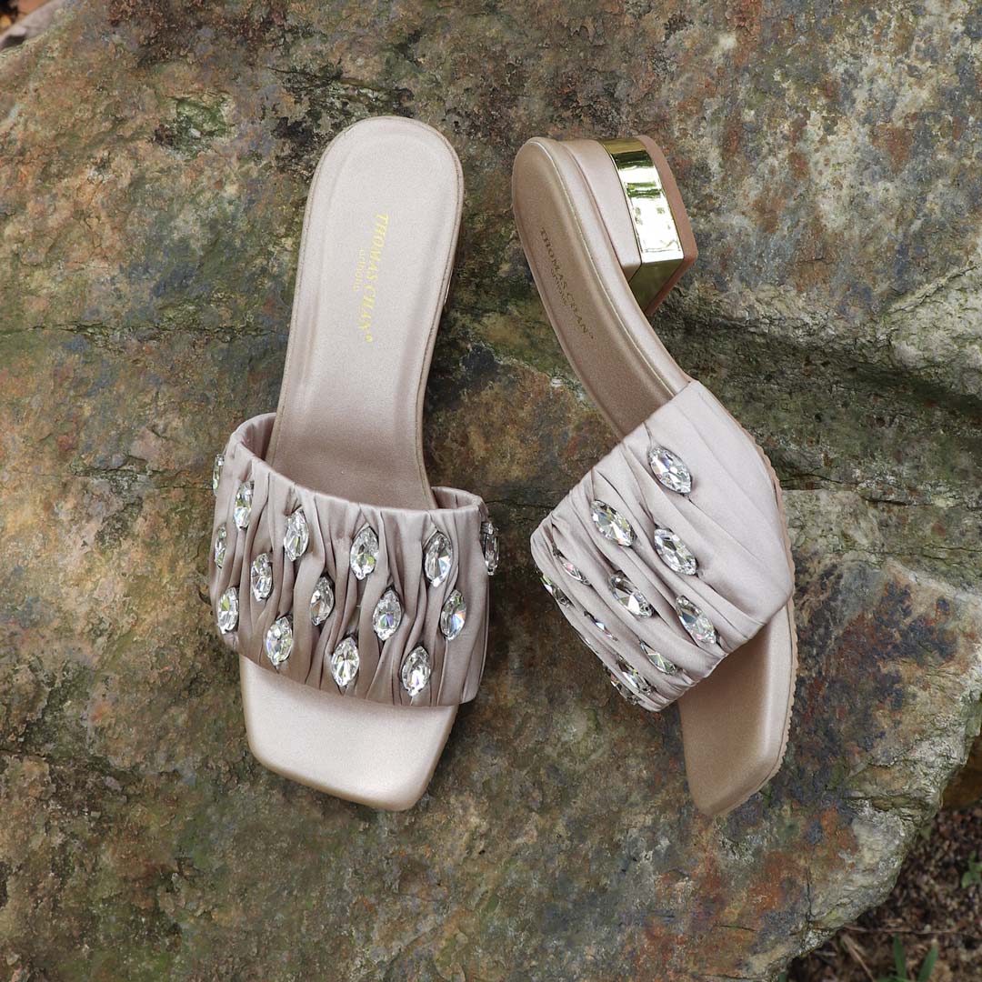 Crystal Lux Satin Evening Heels in rose gold displayed outdoors on a textured stone surface, featuring sparkling crystal embellishments and a glossy block heel — perfect for elegant dinner and evening wear.
