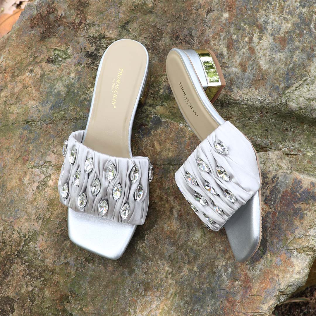 Crystal Lux Satin Evening Heels in silver displayed outdoors on a textured stone surface, featuring sparkling crystal embellishments and a glossy block heel — perfect for elegant dinner and evening wear.
