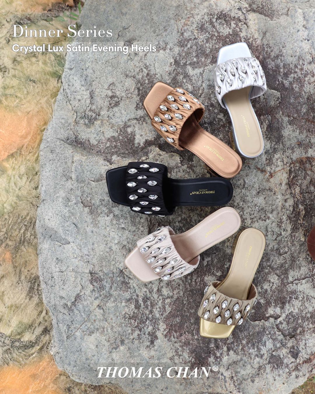 Five pairs of Thomas Chan Crystal Lux Satin Evening Heels in gold, black, rose gold, champagne, and white displayed on a stone surface. Satin ruched straps feature oval crystal embellishments and 4cm block heels, perfect for elegant dinners and formal occasions.
