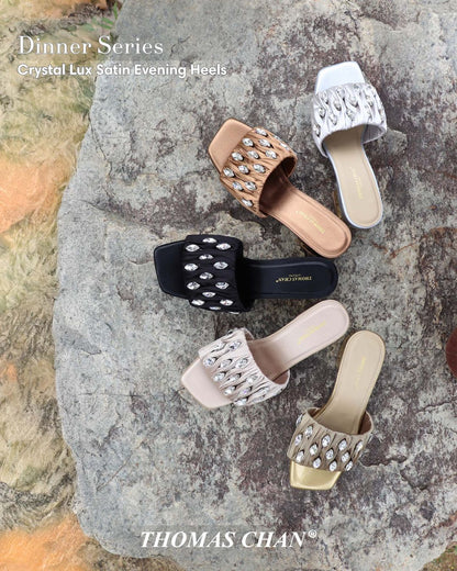 Five pairs of Thomas Chan Crystal Lux Satin Evening Heels in gold, black, rose gold, champagne, and white displayed on a stone surface. Satin ruched straps feature oval crystal embellishments and 4cm block heels, perfect for elegant dinners and formal occasions.