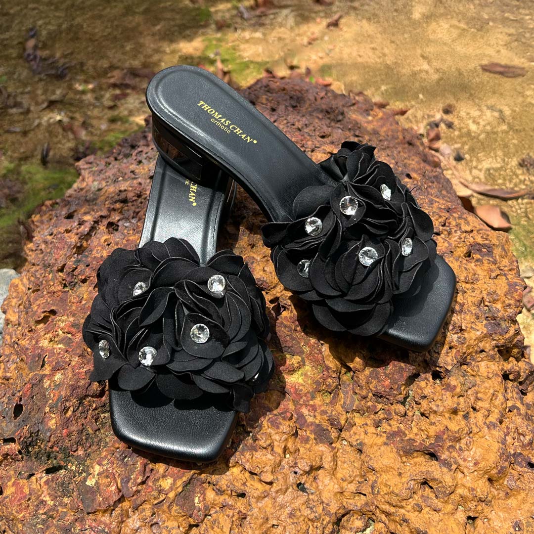 A pair of black evening heels with layered floral embellishments and sparkling rhinestones, set on a low block heel, displayed outdoors on textured rocks.