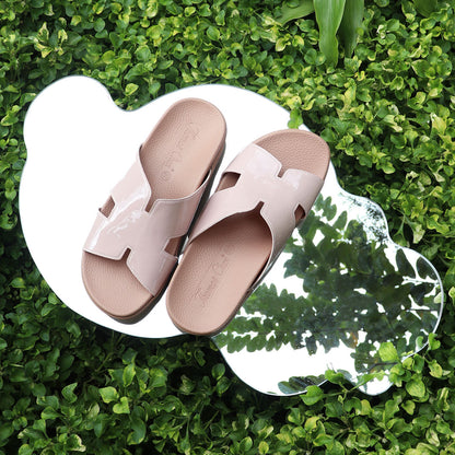A pair of nude pink H-strap orthotic flats from the Thomas Chan Comfy Health Slipper Series is placed on a uniquely shaped mirror, reflecting surrounding green foliage. The glossy finish of the sandals contrasts with the natural background. 
