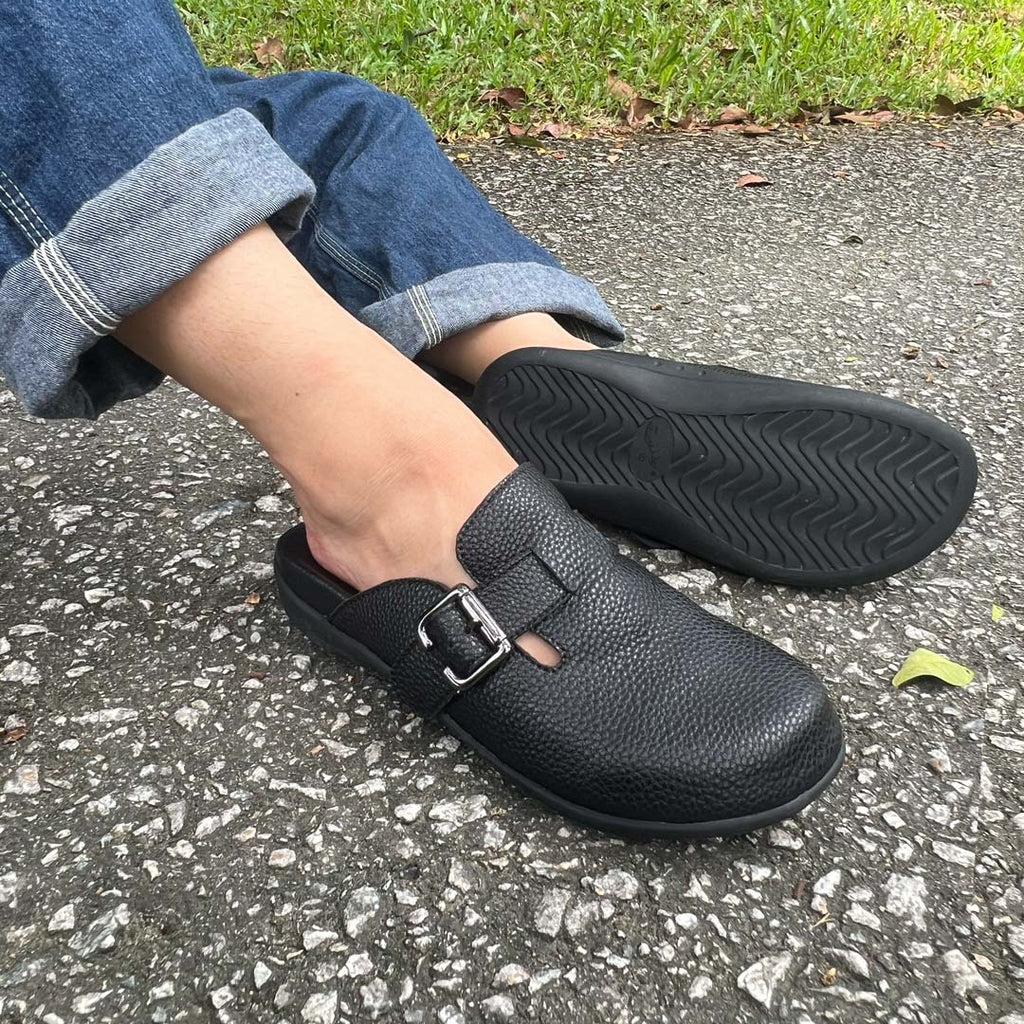 Person wearing rolled-up jeans and a pair of black-coloured Thomas Chan's closed-toe buckle sandals while sitting on a paved surface. The sandals feature a sleek design with a black buckle strap and with arch-support footbed.