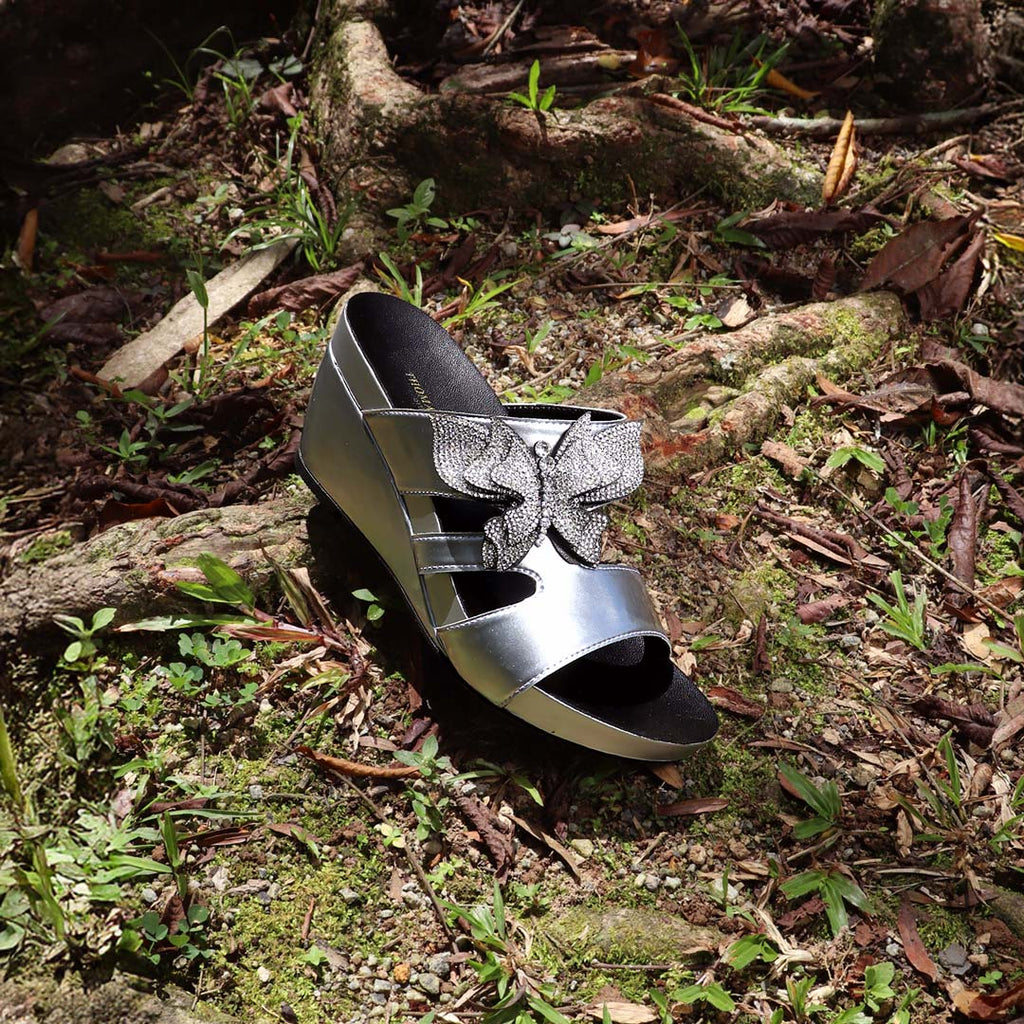 Thomas Chan’s Glimmer Butterfly Dinner Wedges placed on a mossy forest floor with tree roots, dried leaves, and patches of grass, captured in natural daylight. The metallic silver shine and rhinestone butterfly detail contrast beautifully with the earthy, woodland backdrop, evoking a whimsical yet elegant vibe.