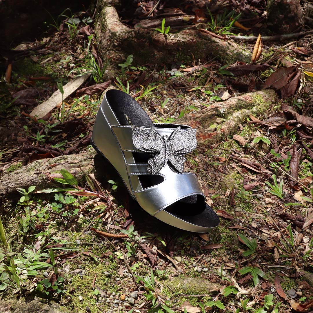 Thomas Chan’s Glimmer Butterfly Dinner Wedges placed on a mossy forest floor with tree roots, dried leaves, and patches of grass, captured in natural daylight. The metallic silver shine and rhinestone butterfly detail contrast beautifully with the earthy, woodland backdrop, evoking a whimsical yet elegant vibe.