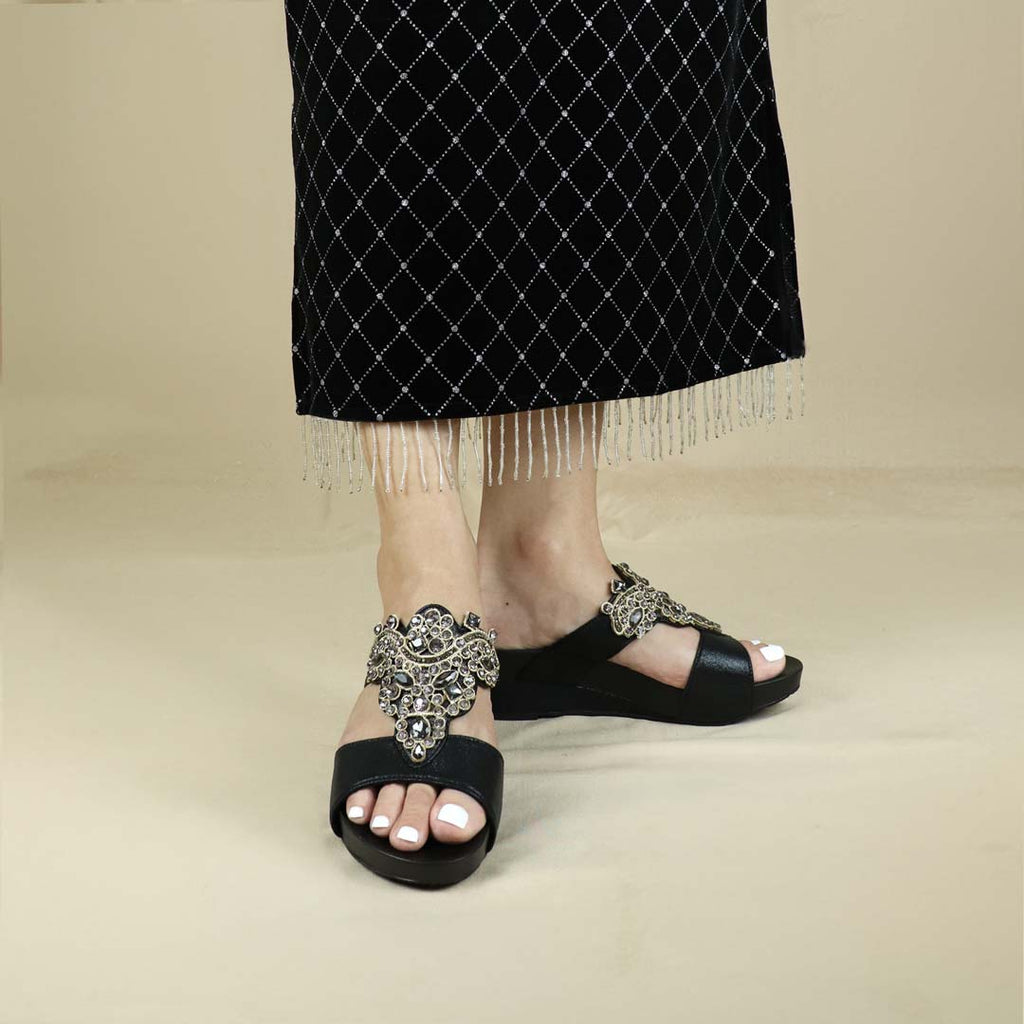 A model showcasing Thomas Chan's Vintage Rhinestone Low Wedge Sandals in black, featuring vintage-inspired rhinestone detailing and an arch-support footbed, standing against a cream backdrop for a refined and elegant presentation.
