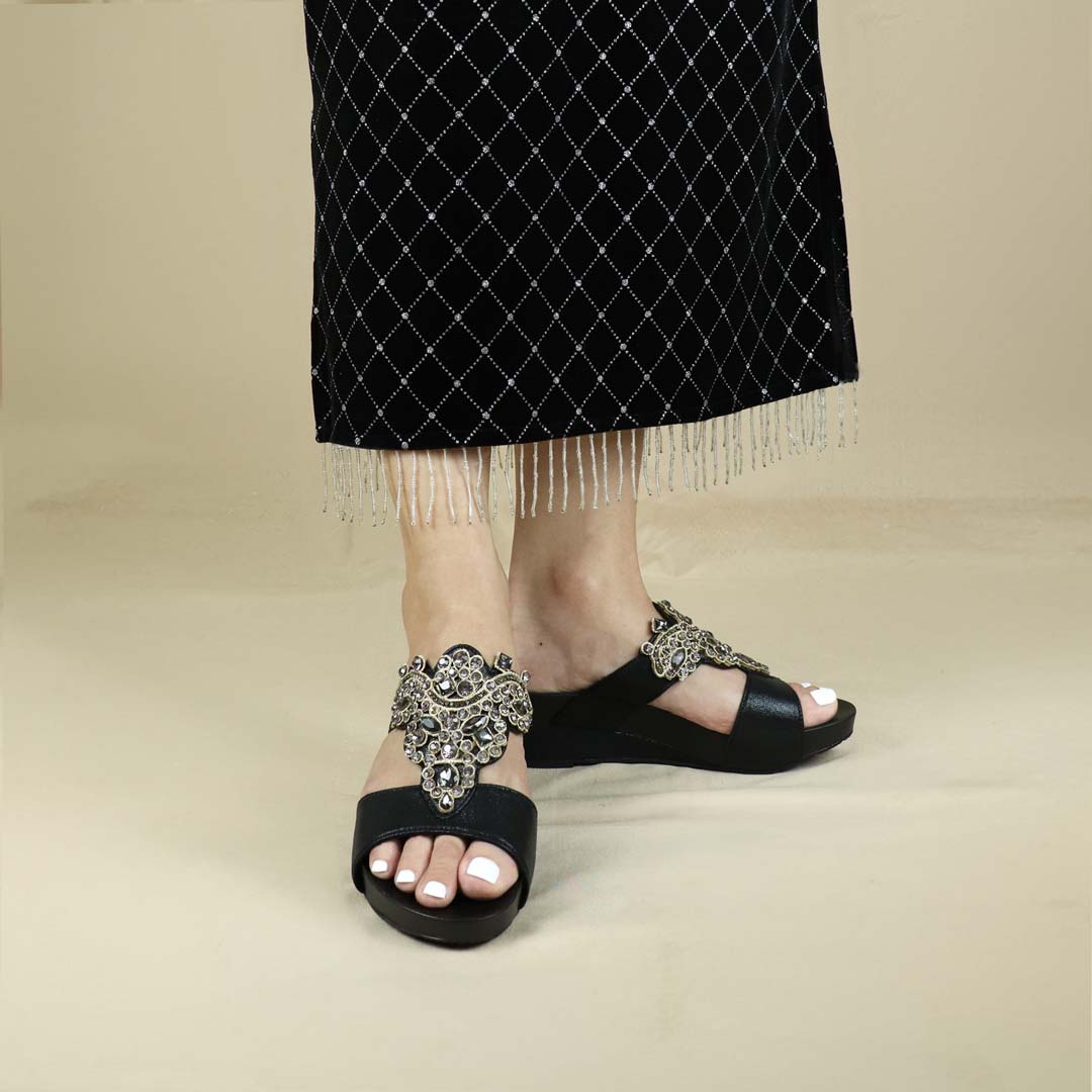 A model showcasing Thomas Chan's Vintage Rhinestone Low Wedge Sandals in black, featuring vintage-inspired rhinestone detailing and an arch-support footbed, standing against a cream backdrop for a refined and elegant presentation.