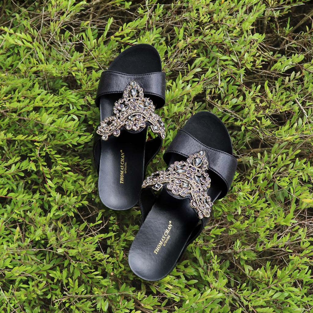 Thomas Chan Vintage Rhinestone Low Wedge Sandals in black, adorned with vintage-inspired rhinestone detailing and crafted with the exclusive Thomas Chan® Orthotic Arch-legance footbed for superior comfort. Set against a backdrop of light green plants, these sandals exude a timeless and sophisticated vibe.