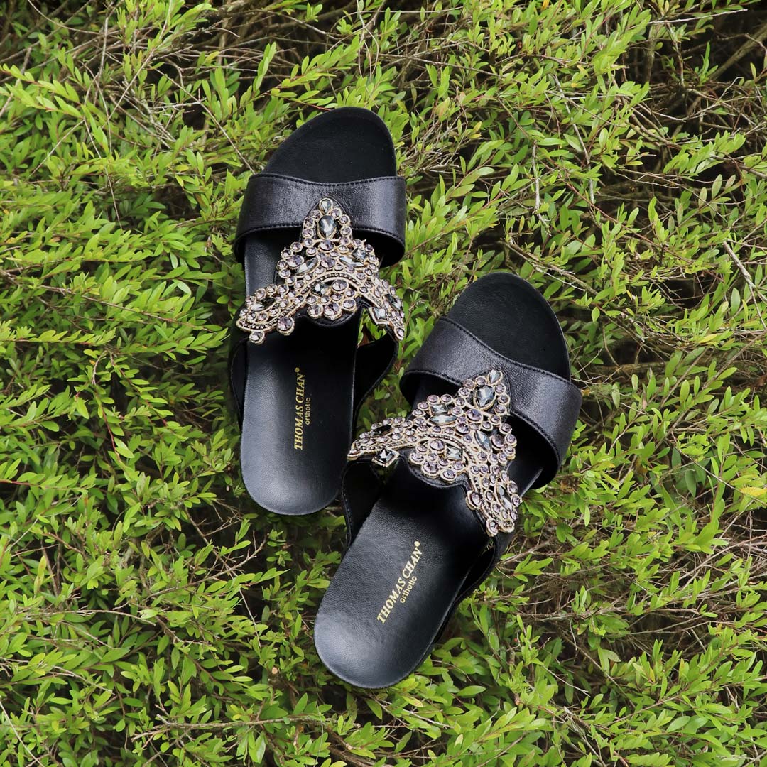 Thomas Chan Vintage Rhinestone Low Wedge Sandals in black, adorned with vintage-inspired rhinestone detailing and crafted with the exclusive Thomas Chan® Orthotic Arch-legance footbed for superior comfort. Set against a backdrop of light green plants, these sandals exude a timeless and sophisticated vibe.