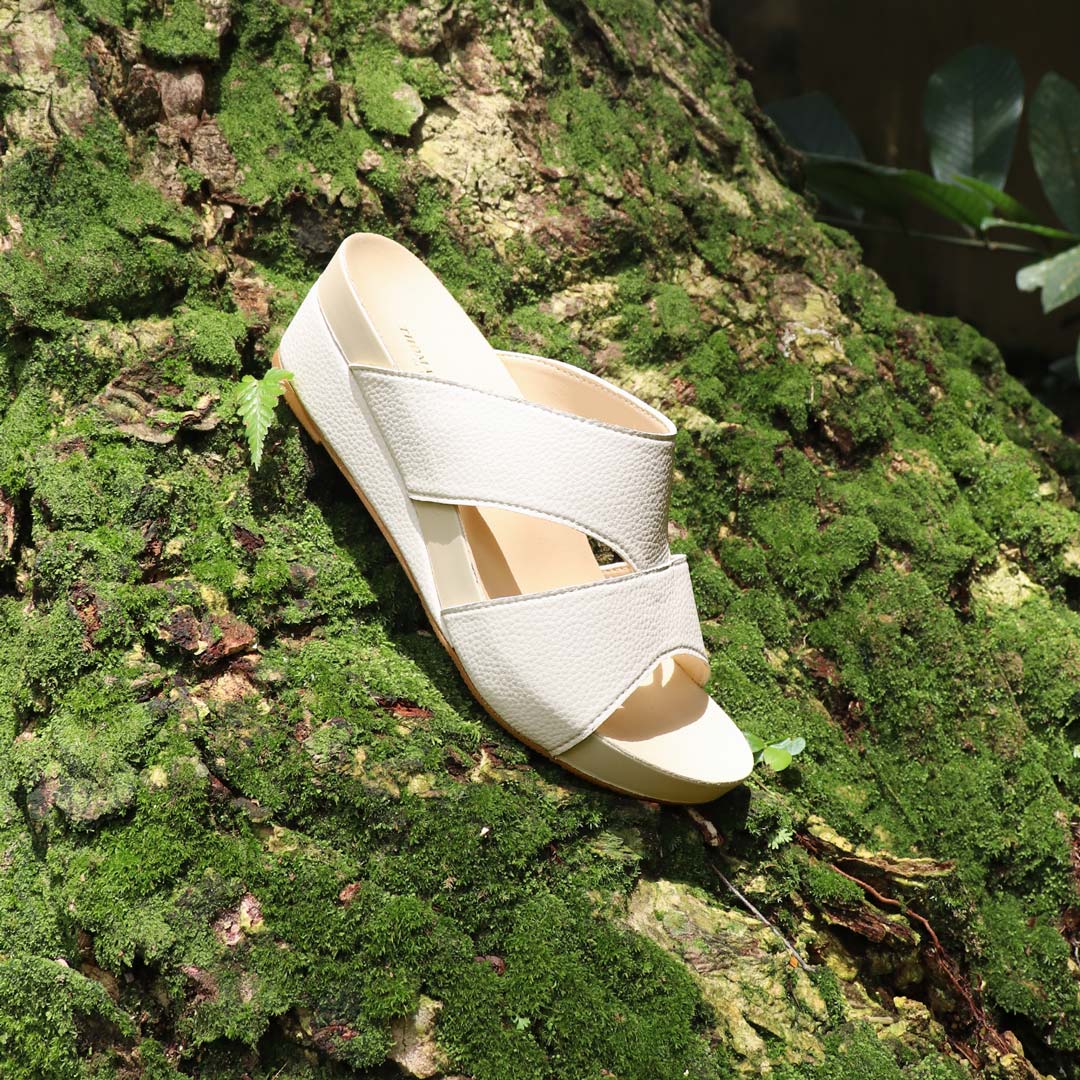 A pair of ivory Pebble Lychee Everyday Sandals from the Thomas Chan ArchLegance Collection, placed elegantly on a moss-covered tree trunk. The sandals feature textured pebbled material and a soft arch-support footbed, blending nature and comfort.