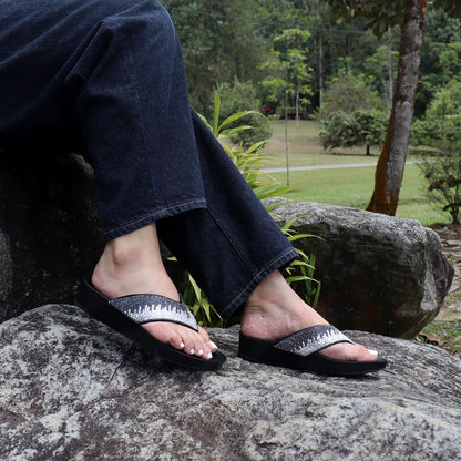 Model seated on a stone outdoors wearing Thomas Chan’s black suede rhinestone flip-flops, highlighting the relaxed style, glittering straps, and comfy flatform sole.