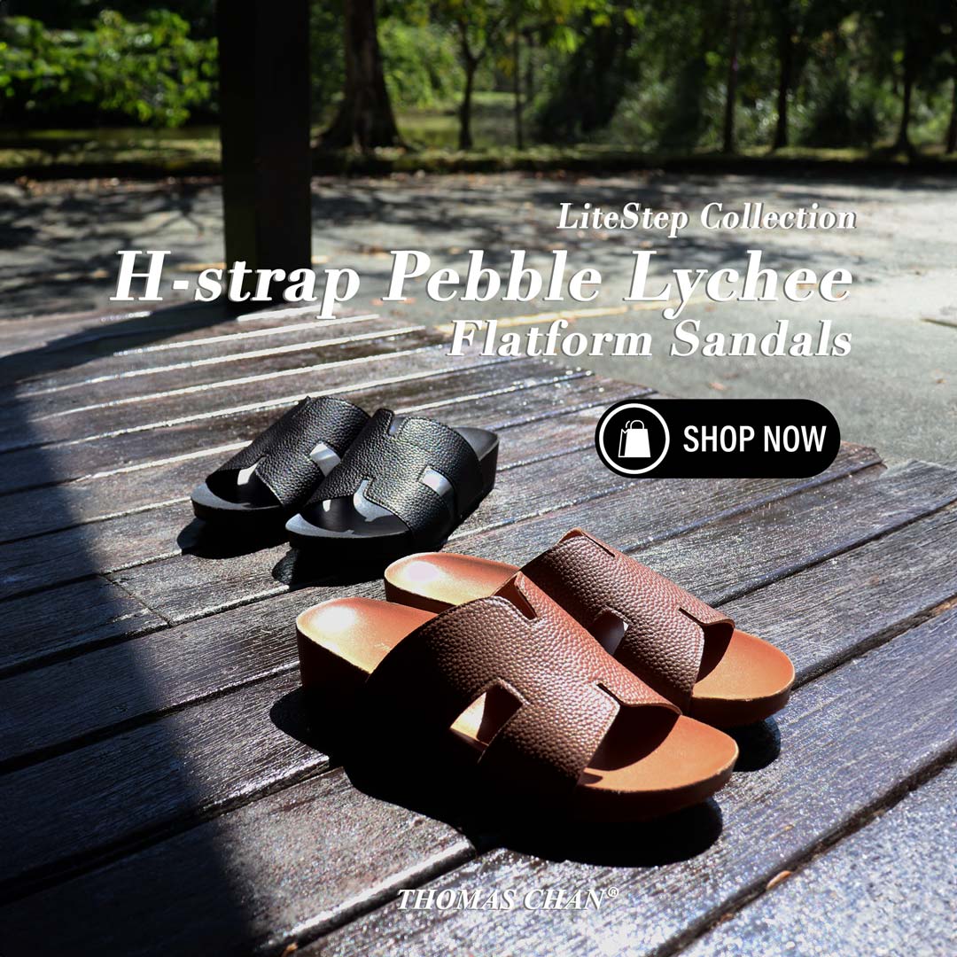 Two pairs of Thomas Chan's H-Strap Pebble Lychee Flatform Sandals displayed on a rustic wooden bench in an outdoor pavilion setting. One pair in rich brown sits in the foreground, showcasing its textured pebble lychee H-strap and flatform sole, while the black pair is positioned slightly behind, creating a contrast against the sunlit wood. The scene is illuminated by natural sunlight, enhancing the sandals’ sleek design and warm earthy tones.