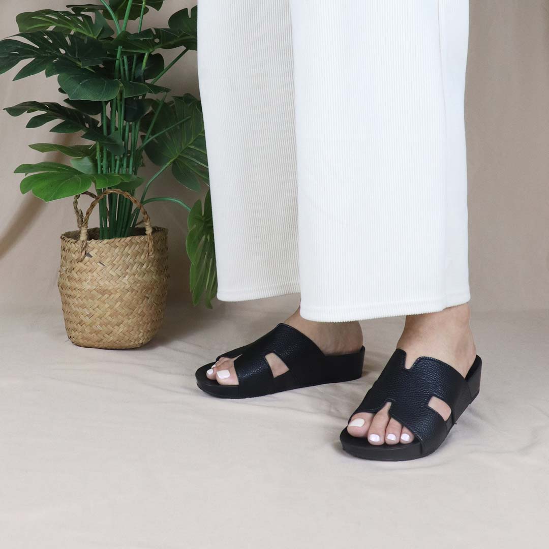 A model elegantly stands wearing Thomas Chan's Black H-Strap Pebble Lychee Flatform Sandals in a studio setting, next to a green plant, showcasing the sleek H-strap design and elevated flatform sole for a chic yet comfortable look.