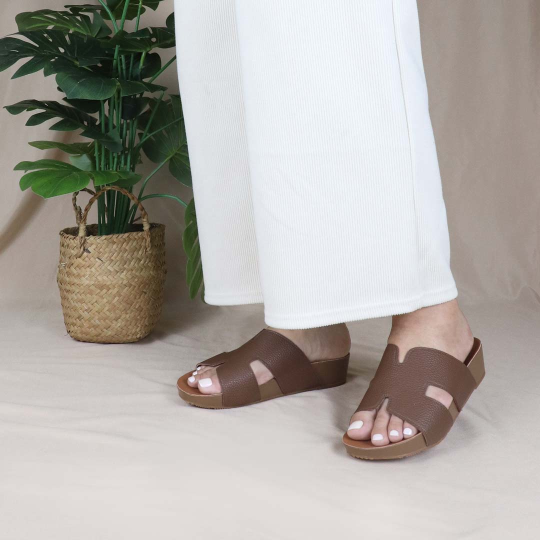 A model elegantly stands wearing Thomas Chan's Brown H-Strap Pebble Lychee Flatform Sandals in a studio setting, next to a green plant, showcasing the sleek H-strap design and elevated flatform sole for a chic yet comfortable look.