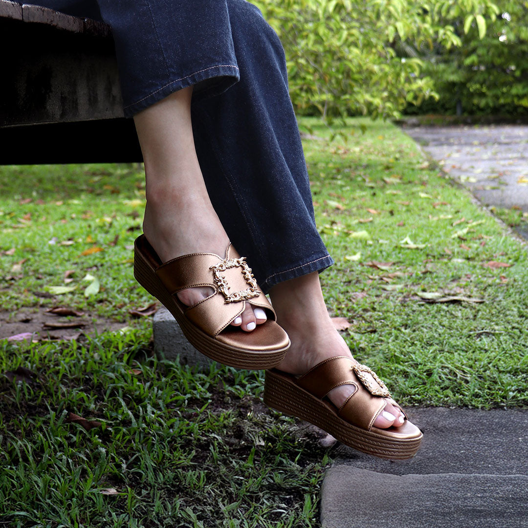 Outdoor model wearing Thomas Chan Vintage Frame Flatform Sandals in bronze, seated casually on a bench. Featuring the bold square buckle design and cushioned footbed from the Cushioned Footbed Series. Captured in a lush garden setting.