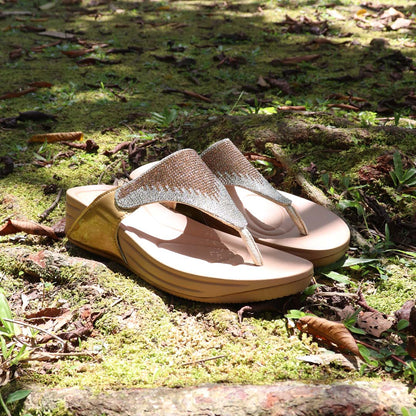 A pair of Gold diamond-embellished flip-flop sandals from the Bounce Care Series is placed on a moss-covered ground with scattered leaves. The sandals feature shimmering crystal details on the straps and a cushioned flatform sole in a neutral beige tone. Sunlight filters through the trees, casting a natural glow on the scene.