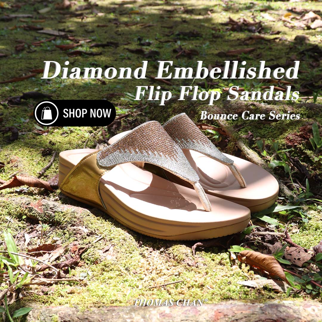 A pair of diamond-embellished flip-flop sandals from the Bounce Care Series is placed on a moss-covered ground with scattered leaves. The sandals feature shimmering crystal details on the straps and a cushioned flatform sole in a neutral beige tone. Sunlight filters through the trees, casting a natural glow on the scene. A 'Shop Now' button overlay is included, along with the text 'Diamond Embellished Flip-Flop Sandals – Bounce Care Series' and the Thomas Chan® logo.