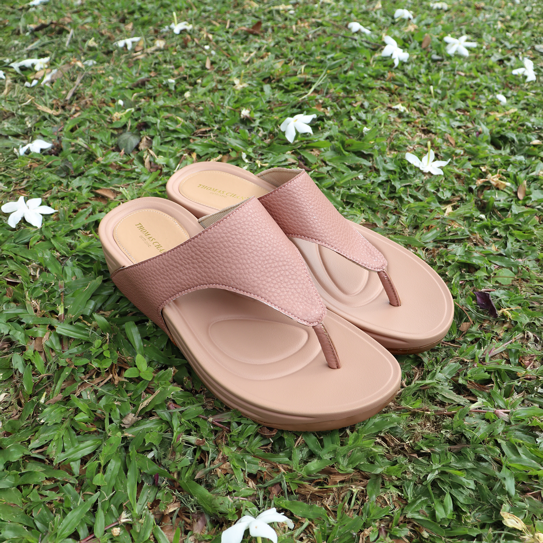 A pair of Thomas Chan’s pastel-tone pink flip-flop sandals with pebble lychee texture resting on a green grass lawn, surrounded by scattered white flowers. The sandals feature a cushioned Bounce Care sole, offering a soft and supportive look in natural outdoor daylight.