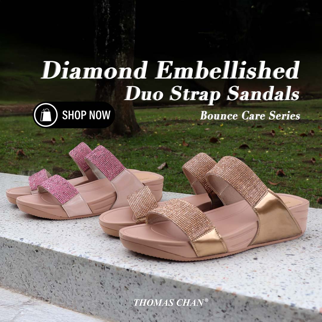 Two pairs of Thomas Chan Diamond Embellished Duo-Strap Sandals from the Bounce Care Series displayed on a stone bench outdoors—one in rose pink and the other in bronze gold. The sandals feature shimmering embellishments and cushioned soles. Text on image reads "Diamond Embellished Duo Strap Sandals – Bounce Care Series" with a 'Shop Now' button and the brand name THOMAS CHAN® at the bottom.